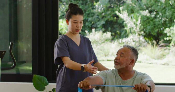Asian therapist in scrubs cueing African American senior man pulling blue band at rehab room window