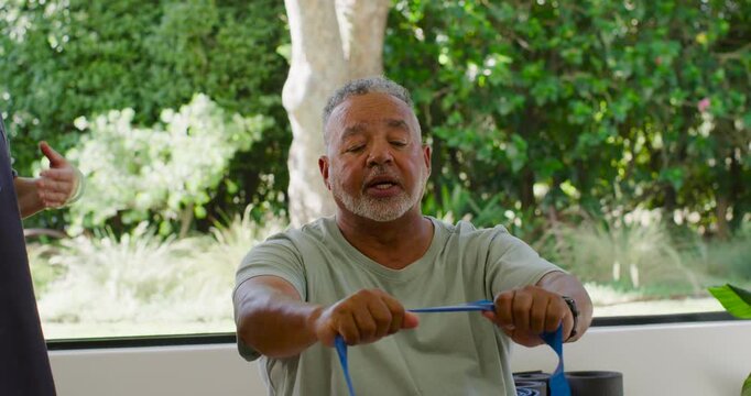 Asian therapist cueing African American mature man pulling blue resistance band for rehab by window