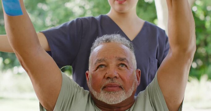 Nurse in scrubs guiding Senior African-American man lifting arms for rehab with blue strap in yard