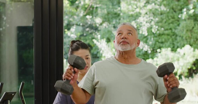 Trainer in scrubs spotting man lifting dumbbells to shoulder, pressing up by glass wall for form