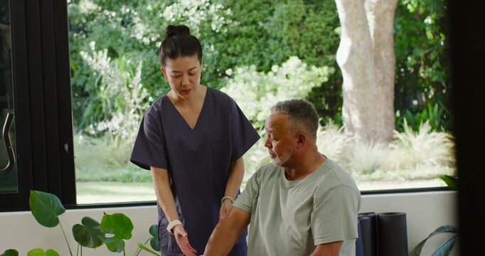 Asian caregiver gesturing, guiding African American senior man lifting hex dumbbell in home rehab