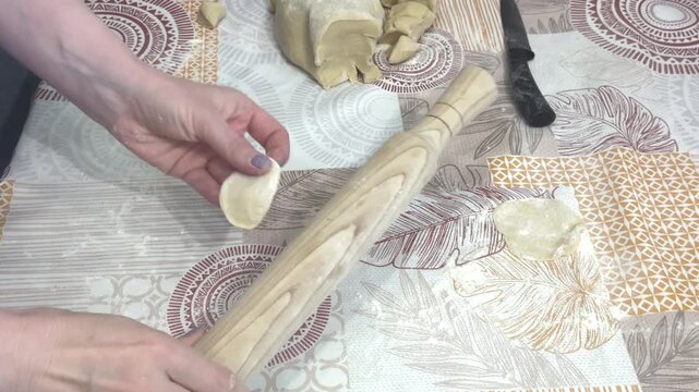 A woman rolls out small pieces of dough with a wooden rolling pin on a patterned tablecloth in a home kitchen, homemade cooking, traditional recipe, culinary process.