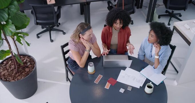 Diverse women, center speaker opening laptop, pointing at sketches at round table, planning design