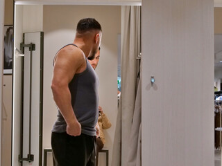 Obraz premium Side view of a muscular man in a grey tank top standing in a brightly lit dressing room. He is looking toward a mirror, which captures his partial reflection. The scene features a minimalist interior 