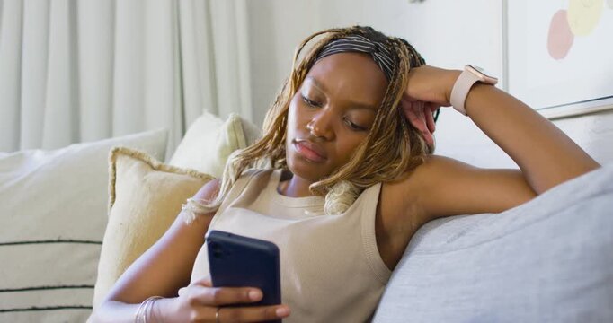 African American woman settling on pillows, scrolling smartphone while resting arm behind head