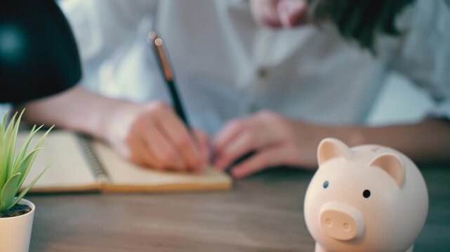 Close-up view of person inserting coin into piggy bank and writing in notebook. Concept of saving money, managing finances, and planning for future, dream achievement, personal finance and success