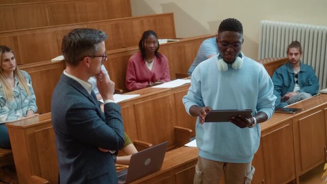 Professor listening to black student presentation during university class while classmates observe. Academic discussion, communication and collaborative learning in modern classroom.
