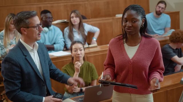 Professor listening to Black female student presentation during university class while classmates observe. Academic discussion, communication and collaborative learning in modern classroom.