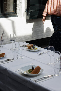 Elegant restaurant table setting with white linen, plates, cutlery and wine glasses in bright natural sunlight. Indoor fine dining scene with waiter, showcasing luxury hospitality atmosphere