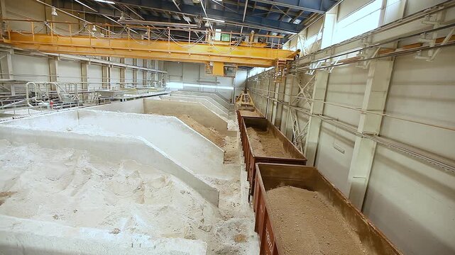 Open freight cars loaded with sand and clay are unloaded in the factory's bulk materials warehouse. Indoors. Raw materials and bulk materials warehouse at a factory producing ceramic tiles