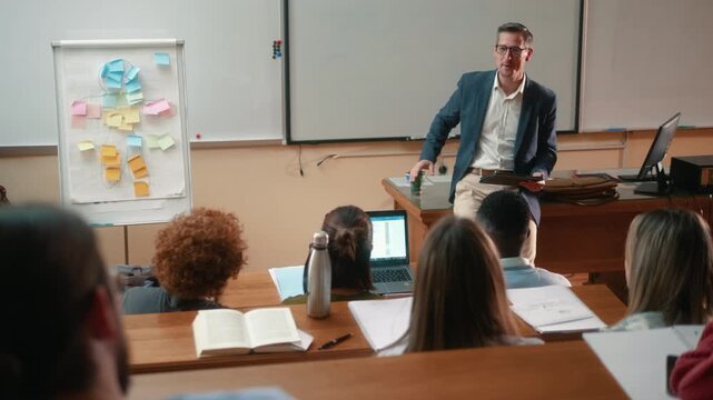 Professor giving lecture and presenting ideas with flipchart and tablet in university classroom. Students listening from behind during academic presentation and discussion.