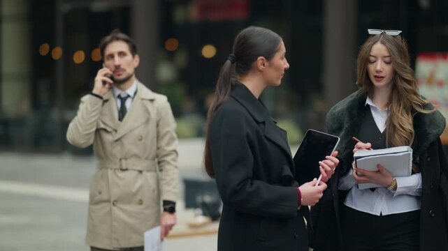 Two businesswomen engage in a professional discussion outdoors while a businessman makes a phone call in the background. Young professionals work and communicate in a dynamic urban setting.