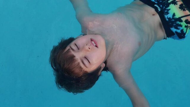 Happy Child relaxing in blue swimming pool. Boy with dark hair floats on his back in clear, bright blue water. His eyes are closed in peaceful relaxation. Serene, calm mood. Tranquility and summer.