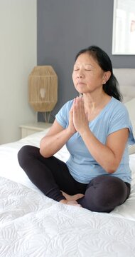 Vertical video: Senior Asian woman meditating cross-legged on bed, breathing, holding prayer hands