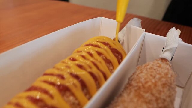 Close-up shot of two freshly prepared, crispy Korean street food corn dogs in a white paper tray. One is being drizzled with a zigzag pattern of bright yellow mustard and red ketchup. The other is coa
