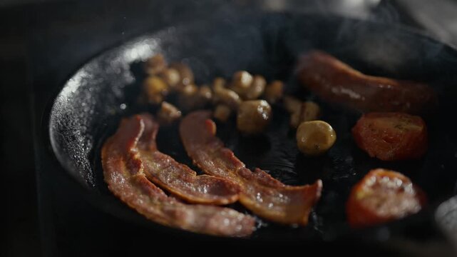 Crispy bacon sizzles in a hot pan with mushrooms. Metal tongs flip bacon, releasing fragrant smoke. Fatty strips curl as golden-brown mushrooms cook beside them