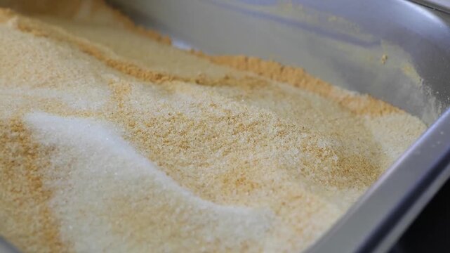 A close-up, high-definition video of various types of bread crumbs, panko, or different shades of flour and bran in a stainless steel professional kitchen container. The granular texture and natural c