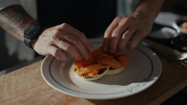 Hands place smoked salmon on soft bread. Focus remains on food preparation, minimal movement. Warm lighting highlights textures and colors. Perfect for cozy breakfast or healthy meal scenes