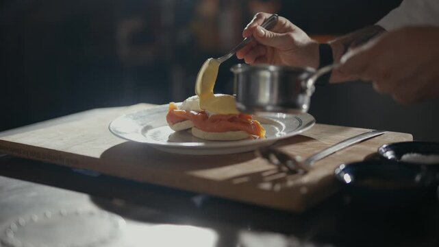 Hands pour custard over eggs and bacon. Dishes sit on a rustic wooden board. Sunlight glows warmly through the kitchen. Chef focuses intently on the dish. Perfect for food or cooking content