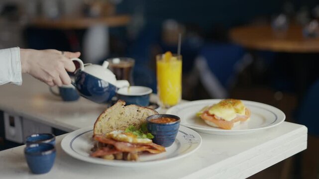 Hand pours tea into a cup. Breakfast plate holds eggs, bacon, and toast. Another plate holds a savory egg dish. Glass of orange juice sits beside the meal. The scene evokes calm, cozy morning dining