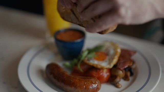 Cozy breakfast scene with sausages, eggs, and toast. Person's hands reach for food with gentle motion. Glass of orange juice adds warm, inviting color. Comforting meal suggests a relaxed morning start