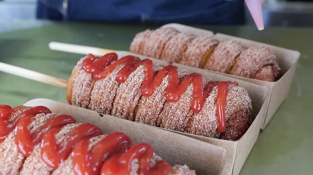Close-up shot of a pastry bag drizzling sweet red sauce over a warm, cinnamon-sugar-coated churro chimney cake on a stick in a paper tray.