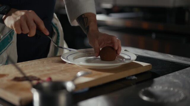 Chef cuts a round food item with a knife. Hand moves carefully over wooden cutting board. White plate sits beside the cutting board. Focus remains on the food preparation process