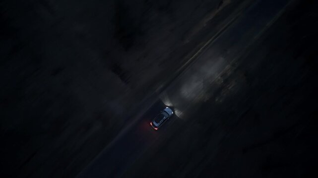 Drone follows a car moving through forest road at night with strong headlights illuminating narrow path in darkness