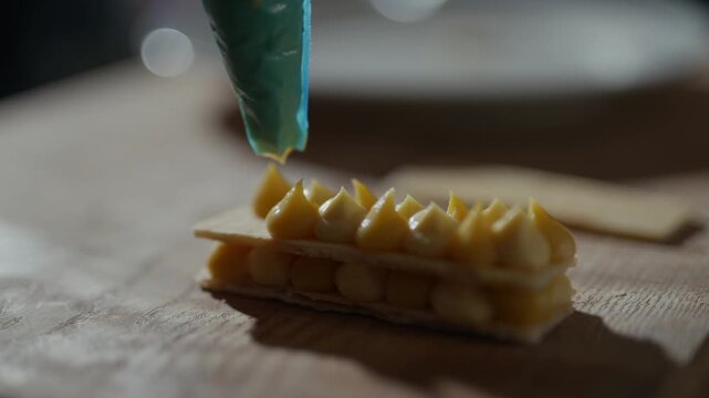 Golden pastry layers gleam under soft light. Blue piping bag releases sweet cream with precision. Wooden surface adds rustic charm to delicate dessert. Perfect for culinary or food-focused projects