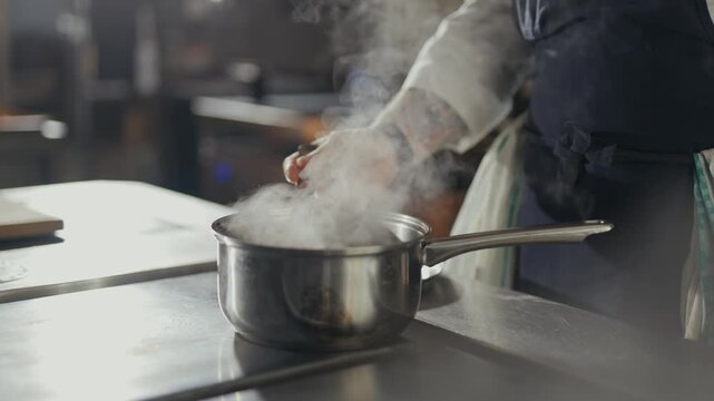 Steam rises from a steaming pot on a stainless steel surface. Chef's hand adjusts a food item inside the pot. The chef's motion suggests precision and care