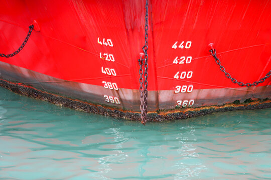 Red ship bow revealing draft marks, hull, anchor chain, and waterline with barnacles, showing maritime industry concepts, shipping, transportation, weight, and history