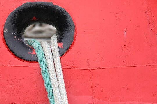 Red boat hull featuring a weathered black hawsehole, securely holding two thick, braided mooring ropes, highlighting maritime details and textured surfaces
