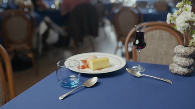 Blue tablecloth holds dessert and wine. Silverware gleams beside a slice of cake. Empty chairs suggest a quiet, intimate meal. Soft lighting hints at relaxed, elegant dining