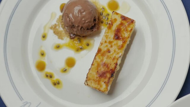 Warm dessert sits on a white plate. Sauce drips around a scoop of ice cream. Golden-brown cake slice adds contrast. Passion fruit seeds garnish the dish. The plate features elegant blue rim details