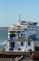 Fototapeta premium A large passenger and vehicle ferry, the Victoria of Wight, navigates the water near a quaint white house with blue window frames in Portsmouth Harbour UK