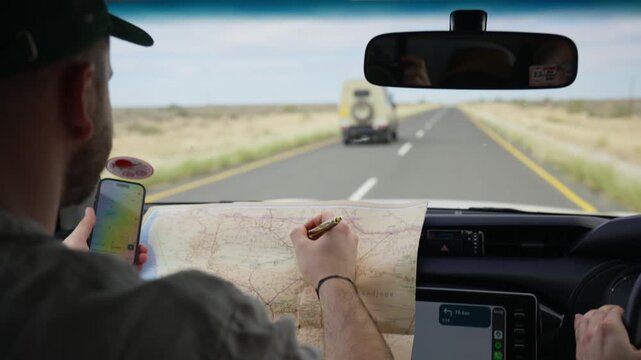 Adventurous man planning a road trip, marking destinations on a paper map of Namibia, Africa while using smartphone for navigation inside a moving safari car