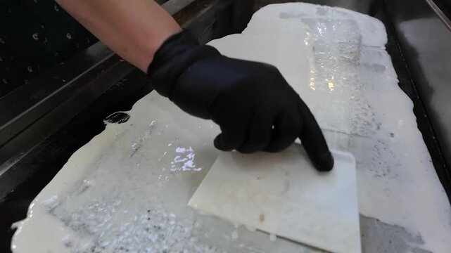 A close-up shot of a chef's hand in a black glove using a spatula to spread white batter on a hot, flat griddle to prepare a popular South Indian or Malaysian dish. The process involves skilled spread
