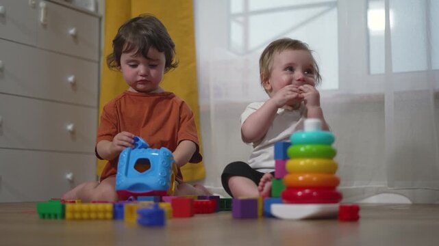 Two toddlers playing with colorful toys on floor. Children sit together with blocks and stacking rings. Kids play indoor educational game. Toddlers learn with toys. Children develop skills.