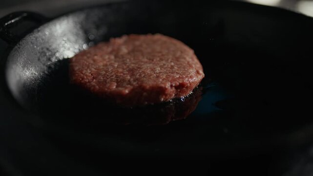 Sizzling patty cooks in a black skillet. Dark pan glows under gentle heat. Juices bubble around the meat. Cooking process feels warm and inviting. Perfect for food and culinary content