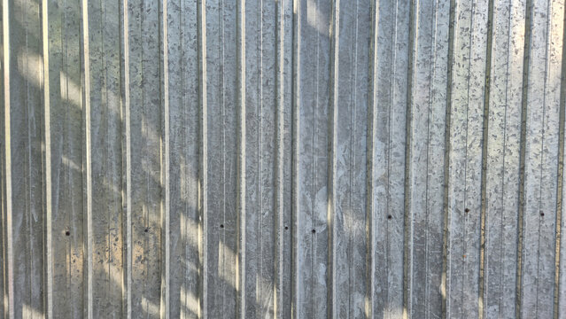 A close-up view of a vertical corrugated galvanized steel fence, showing metallic textures and dappled shadows from nearby foliage.