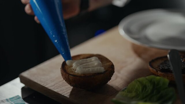 Close-up of hand squeezing cream into burger. Fingers hold blue piping bag gently. Cream swirls onto toasted bun base. Food prep scene feels calm and precise