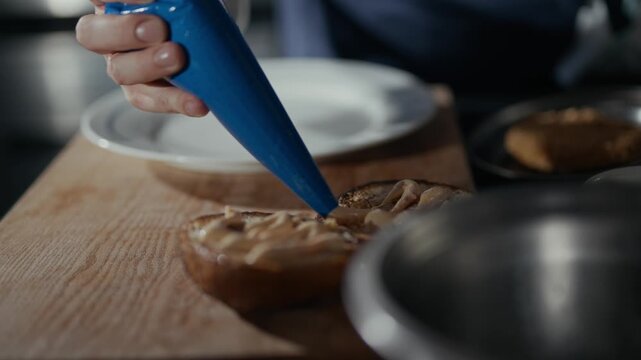 Hand squeezes creamy filling onto toasted bread. Close-up shows careful, precise action. Wooden board, metal bowl, and white plate create simple backdrop
