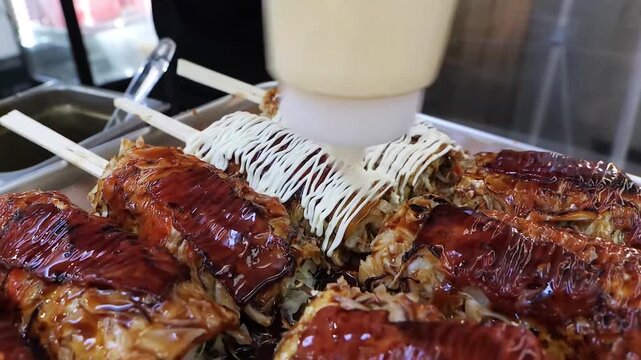 A close-up, high-angle shot captures a vendor drizzling white Japanese mayonnaise from a squeeze bottle onto several pieces of freshly prepared okonomiyaki (hashimaki) on wooden skewers. The savory pa