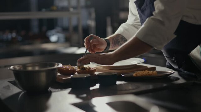 Dramatic kitchen light highlights chef's focused hands. Chef delicately garnishes dish with precision and care. Professional kitchen ambiance suggests high-end culinary craft