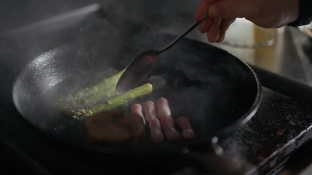 Sizzling pan holds meat, asparagus, and sausages. Steam rises as condiment drips from a hand. Cooking scene exudes warmth and savory appeal. Ideal for culinary, restaurant, or food prep visuals