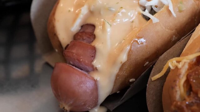 A macro, close-up shot of a gourmet hot dog served in a soft, toasted bun. The hot dog is generously topped with a rich, creamy savory sauce (like a spicy mayo or signature dressing) and garnished wit