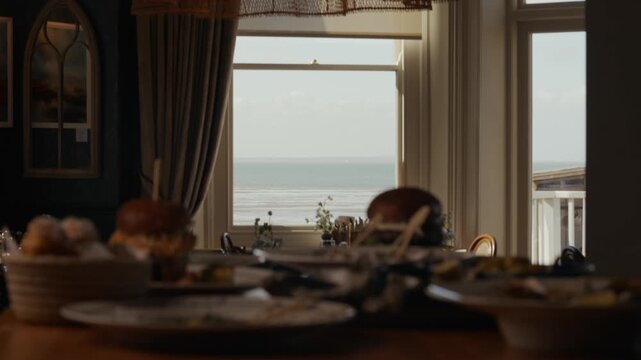Dinner table cluttered with plates and food. Large window frames sea view and distant horizon. Soft light bathes the room. No people visible in this shot