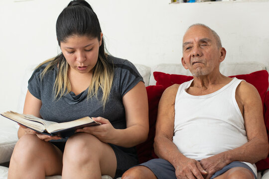 Granddaughter reading book aloud to visually impaired grandfather sitting together on couch