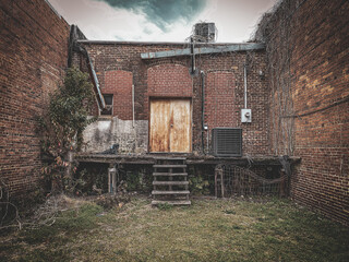 An old brick building with weathered wooden doors sits between two walls with overgrown vines rusted metal pipes and a patchy grassy yard beneath a cloudy sky in an urban alley © DrewDuzz