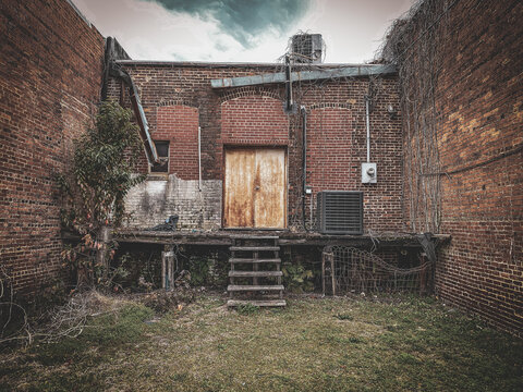 An old brick building with weathered wooden doors sits between two walls with overgrown vines rusted metal pipes and a patchy grassy yard beneath a cloudy sky in an urban alley